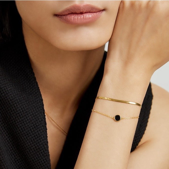 Close-up of a woman wearing a gold bracelet with a black circle charm and pearl earrings.