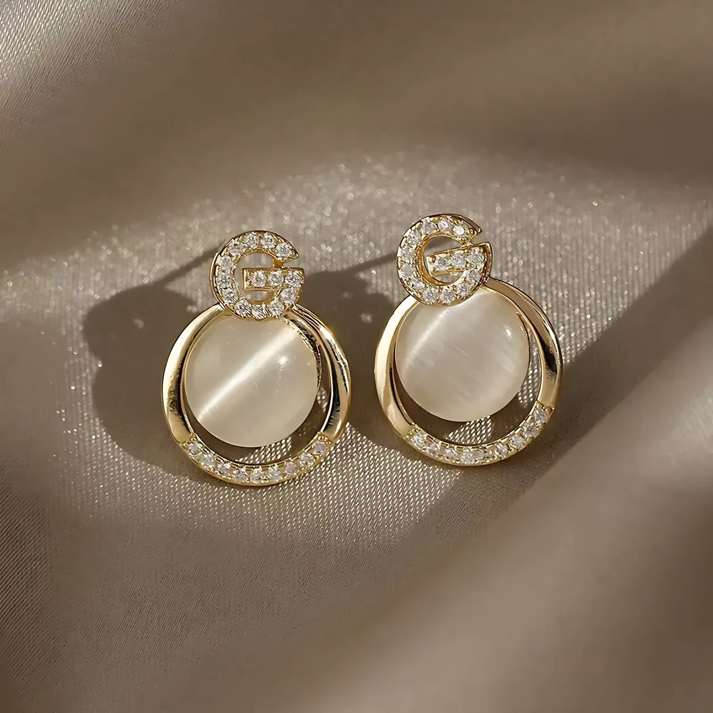 Gold hoop earrings with white opal-like stones and the letter "G" outlined in crystals.