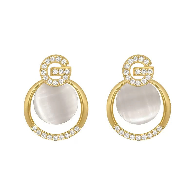 Gold hoop earrings with white opal-like stones and the letter "G" outlined in crystals.