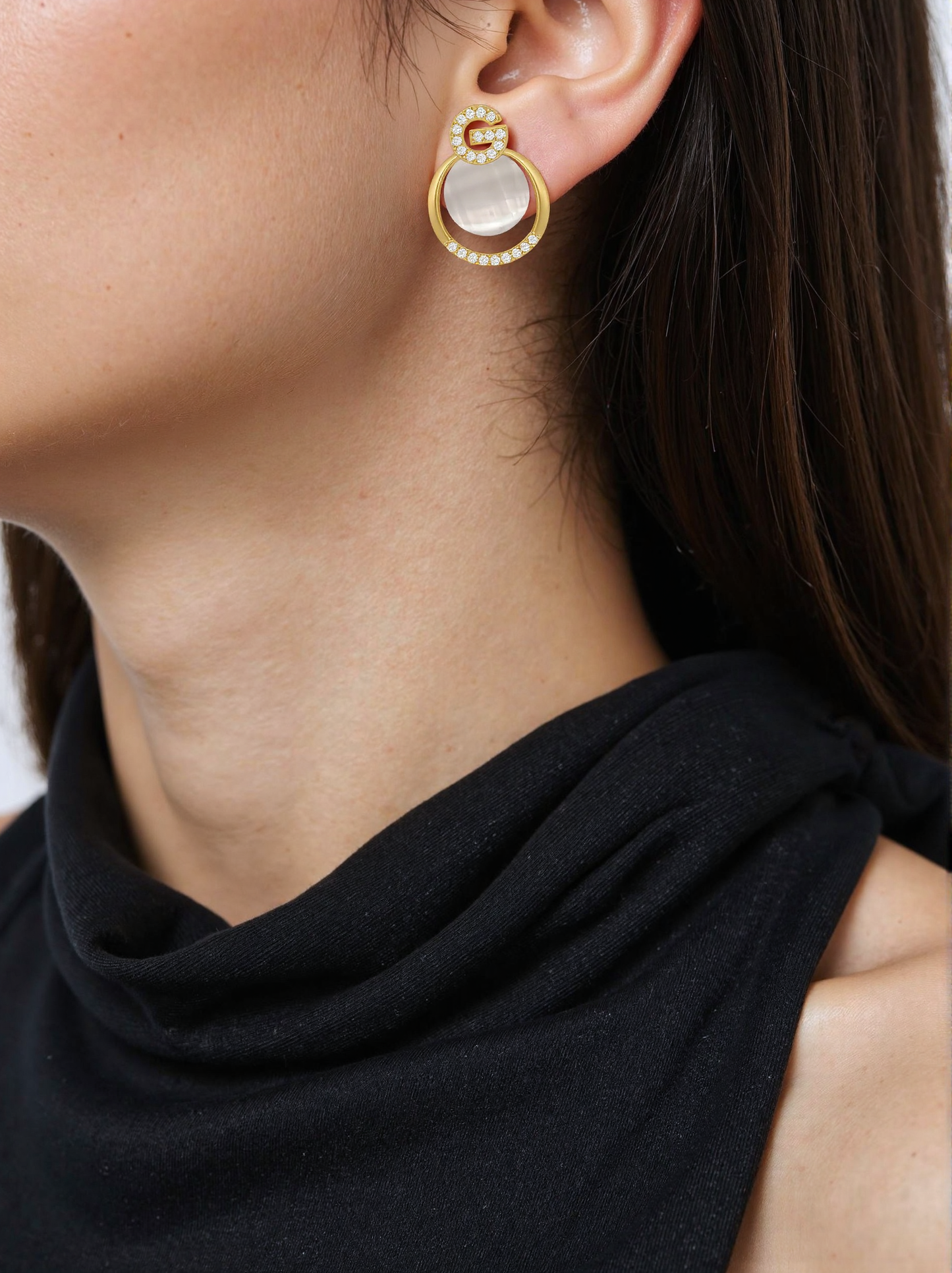 Close up of a woman wearing gold hoop earrings with white opal-like stones and the letter "G" outlined in crystals.