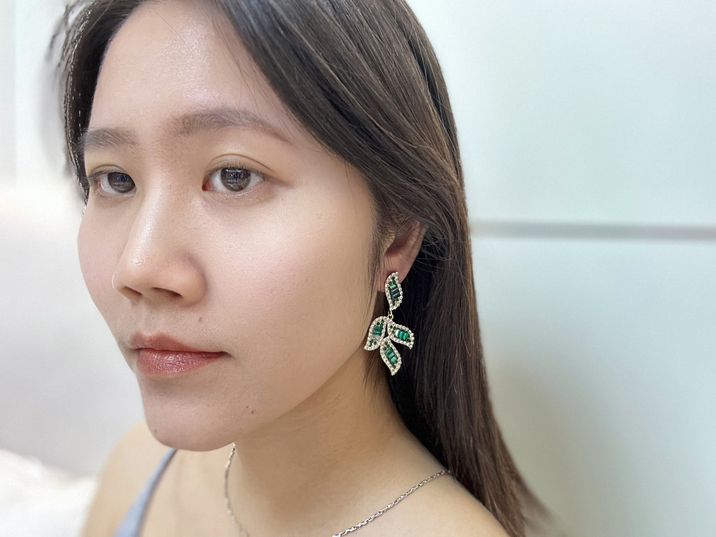 Close-up of a woman wearing gold-toned earrings featuring leaf-shaped designs adorned with baguette-cut green crystals and white cubic zirconia.