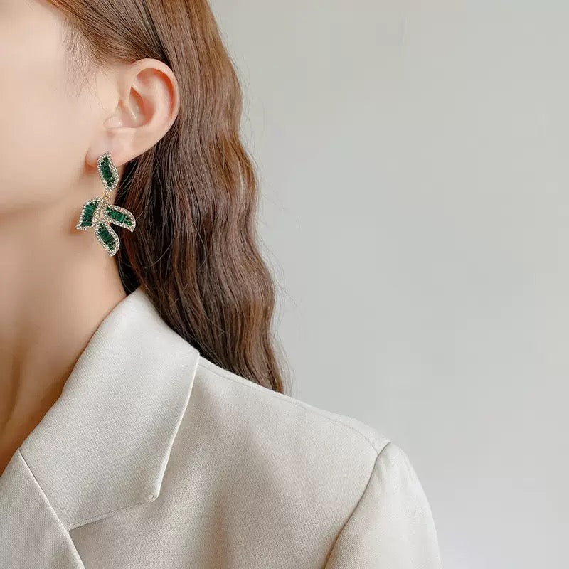 Close-up of a woman wearing gold-toned earrings featuring leaf-shaped designs adorned with baguette-cut green crystals and white cubic zirconia.