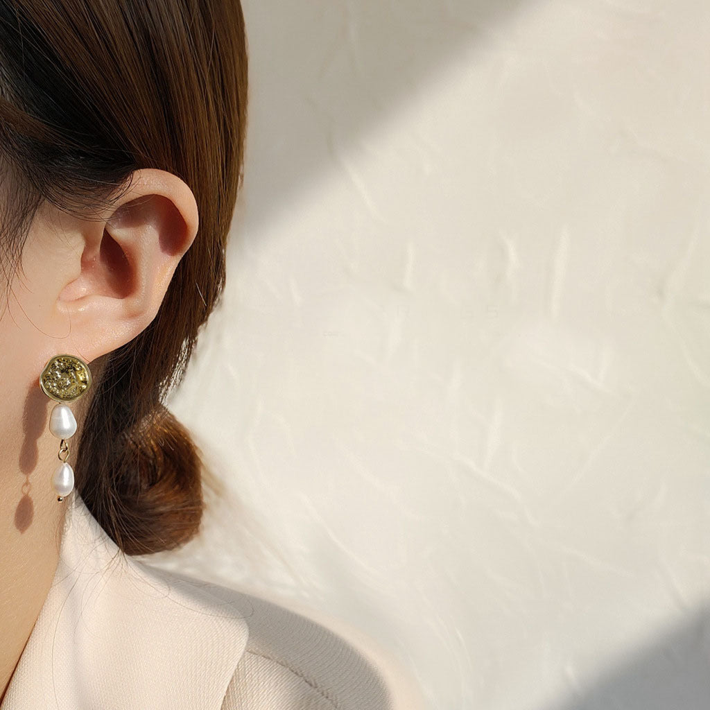 Close-up of a woman wearing a pair of gold-toned earrings with textured discs and pearl drops.